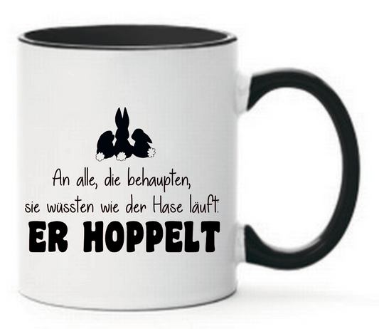 Schwarze Tasse mit Spruch An alle, die behaupten, sie wüssten wie der Hase läuft. Er hoppelt und drei Comic Hasen auf der Tasse